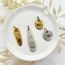 An overhead shot of three organic, hand-stamped charms resting on a white ceramic dish. The charms feature a hammered, pebble-like texture in gold and silver finishes, each stamped with "Gigi" in a delicate script. They include a long oval, a teardrop shape, and a small round disc. The scene is styled with a neutral bohemian aesthetic, featuring soft dried botanicals and earthy tones, highlighting the artisan, handcrafted quality of the jewelry.