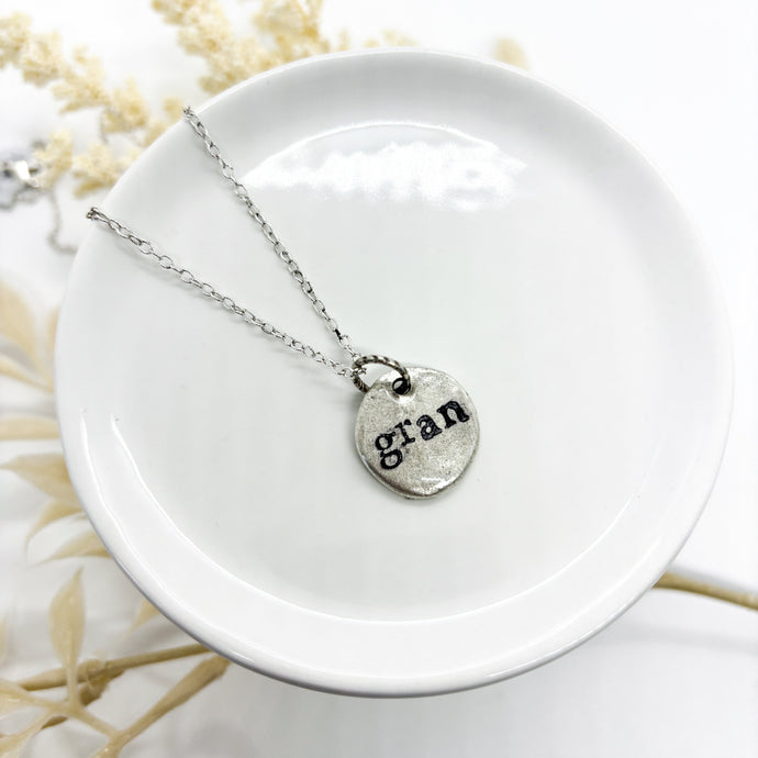 Silver necklace with a round pendant and gran ink stamped & sealed with resin on a white dish with dried flowers in the background