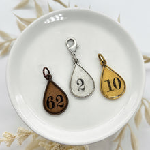 Teardrop-shaped number charms displayed on a white dish. Three metal finishes are shown: antique silver with the number 2, rustic copper with the number 62, and warm gold with the number 10. Each charm has a stamped number and a coordinating loop ready to be added to a necklace, bracelet, journal & keychain. Neutral dried botanical stems surround the dish, creating a soft, minimal background that highlights the personalized charm options for custom jewelry designs or meaningful keepsake gifts.