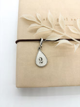 mage featuring a tear-shaped metal charm clasped neatly on a journal with a lobster clasp. The charm is a silver finish and displays an ink stamped number 2 sealed beneath a layer of resin finish.