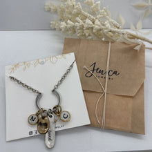 An unedited picture of product packaging set displaying a silver charm necklace mounted on a white display card next to a kraft paper gift envelope tied with string and branded "Jenica Jewelry". The necklace features a horseshoe link holding initial charms "N" and "C", an oval charm, and an elongated teardrop pendant on a silver chain.