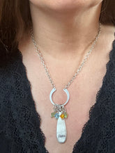 Close-up of a person wearing a silver chain necklace styled with a horseshoe-shaped link pendant attached to the chain. Suspended from the horseshoe are several small, colorful birthstone crystal charms and an elongated, flat teardrop-shaped silver pendant hand-stamped with the name "Nana" near the bottom. The piece is worn over a black lace V-neck top.
