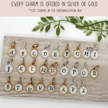 Product graphic titled "EVERY CHARM IS OFFERED IN SILVER OR GOLD" with the note "LIST CHARMS IN THE PERSONALIZATION BOX". Below, a grid displays circular initial pendant charms for letters A through Z on a textured stone slab with green botanical leaves behind it. Each small round pendant has a smooth white surface, a stamped uppercase letter, and a twisted jump ring in either gold or silver metal finish