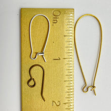 A top-down close-up displays three styles of jewelry ear wire hooks arranged next to a brass ruler for scale. On the left side of the ruler, a silver kidney-style ear wire measures just over 1 inch long, and an antiqued bronze-tone French hook sits below it. On the right side of the ruler, a long, dramatic gold kidney-style ear wire extends to approximately 2 inches in length. The flat-lay layout highlights the size and shape variations available.