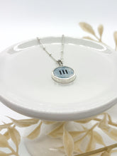 A product photograph features a sterling silver satellite chain necklace resting on a small, glossy white ceramic display stand. The round pendant has a thick silver bezel framing a light blue watercolor background with the angel number "111" printed in black. The background is clean and white, accented by delicate, faux white leaf sprigs surrounding the base of the stand.
