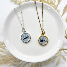 Round resin-sealed 666 angel number necklaces in gold and silver settings with a blueish background. 666 represents letting go of the past and to focus on the present.