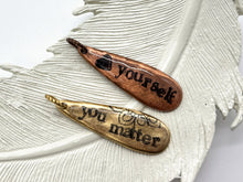 Two rustic, teardrop-shaped metal pendants rest on a textured white decorative feather. On the left, a copper charm is hand-stamped with a black heart icon and the vertical text "love yourself." On the right, a gold brass charm features the phrase "you matter" alongside a delicate floral flourish. Both charms have an antiqued finish with darkened lettering and are topped with small, twisted metal jump rings.