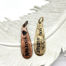Two rustic, teardrop-shaped metal pendants rest on a textured white decorative feather. On the left, a copper charm is hand-stamped with a black heart icon and the vertical text "love yourself." On the right, a vintage gold brass charm features the phrase "you matter" alongside a delicate floral flourish. Both charms have an antiqued finish with black lettering and are topped with small, twisted metal jump rings.