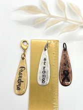 Three hand-stamped metal charms shown alongside a gold ruler for size reference, measuring approximately 1.75 inches long. From left to right: a gold "Grandma" charm with a lobster clasp attachment, a silver "strong" charm with a jump ring, and a copper charm featuring a stamped cancer awareness ribbon. The image demonstrates different metal finishes and attachment options for jewelry or journals.
