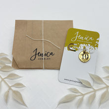 Soft beige botanical accents surround beautiful packaging for the lucky number charm, adding a neutral, organic touch. The numeric charm is tied on a beautiful white and metallic gold card with Jenica Jewelry featured at the top while resting on its top layer of packing which is a recyclable kraft paper bag with Jenica Jewelry's logo stamped on the front & tied off with a simple and delicate white string