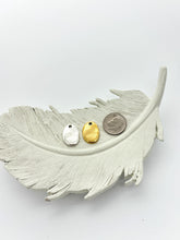 A wide shot of two oval jewelry charms and a United States dime resting on a large, white decorative feather. The charms—one silver and one gold—are shown from the back, displaying a smooth, slightly textured surface with a small hole at the top. The dime is placed to the right of the charms to provide a scale for their size. The entire arrangement is set against a plain white background.