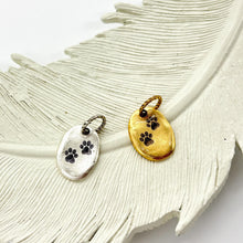 Showing two small, oval-shaped jewelry charms resting on a large, white decorative feather. The charms feature two tiny, dark paw prints stamped into the center. One charm is made of polished silver, while the other is a warm, brushed gold. Both charms have a twisted metal jump ring at the top. The background is a clean, bright white.