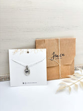 A silver necklace with an oval charm displayed on a white branded jewelry card. Next to the card is a small brown kraft paper envelope tied with white string, featuring the "Jenica Jewelry" logo. The arrangement is set against a white wooden background with a decorative white leaf in the foreground.