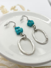 A pair of boho-chic dangle earrings featuring three stacked turquoise stone chips and a large, hammered silver oval pendant. The earrings are resting at an angle on a white ceramic jewelry dish, showcasing the organic texture of the metal and the vibrant blue of the natural stones.