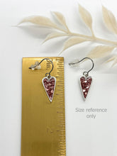 Text: A pair of handcrafted heart-shaped drop earrings shown next to a brass ruler to demonstrate their small, delicate size. Shows they measure a little over 1" in length