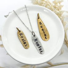 Three hand-stamped metal pendants featuring the word "Resist" in a vintage typewriter font. Two pendants are antiqued gold brass and one is polished silver on a delicate chain. They are displayed on a white ceramic jewelry dish with a soft-focus floral background. The metal has an organic, hammered, and slightly irregular oblong shape.