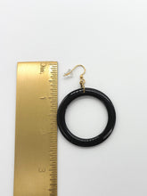 A top-down product photo on a clean white background showing a single statement earring positioned next to a gold metal ruler for scale. The earring features a large, glossy black resin hoop hanging from a gold-toned fishhook wire with a clear rubber stopper. Two small gold jump rings, one with a twisted texture, connect the hoop to the wire. The ruler indicates the black hoop is approximately 1.5 inches in diameter, while the total drop length of the earring is about 2.5 inches.