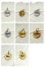 A grid of eight photos features small, organic-shaped metal charms with hand-stamped names, each resting on a white, feathered background. The charms come in silver or gold finishes with textured, twisted jump rings. Stamped names include "Gma," "nan," "nina," "nam," "gran," and "nona" in a typewriter-style font, plus "Nana" and "Mom" in a cursive script. Each pendant has a rustic, hammered appearance with blackened