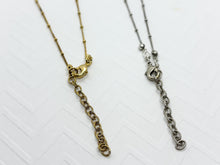 Two delicate satellite necklaces, one gold and one silver, are shown from above on a white, textured chevron background. Both feature thin chains with tiny metallic beads. The focus is on the center, showing lobster claw clasps hooked into textured, round-link extender chains. The gold necklace is on the left with a polished finish, and the silver necklace is on the right with an antiqued sheen.