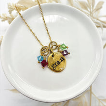 A top-down, high-angle studio shot of a personalized gold-toned necklace resting on a small, round white ceramic dish. The necklace features a dainty cable chain and a rustic, organic-shaped gold circular pendant hand-stamped with the word "gran" in a lowercase serif font. Hanging from decorative rope-textured rings are four faceted bicone crystal charms in light blue, ruby red, peridot green, and amethyst purple. Dried white floral stems softly blur in the bright, clean background.