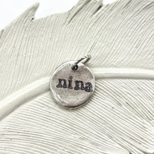 A small, round fine silver plated pendant with a rustic, organic "pebble" shape and a high-shine hammered texture. The name "NINA" is hand-stamped in a clean, minimalist lowercase font in the center. The charm is attached to a sturdy silver jump ring and displayed against a soft, neutral background, highlighting its handcrafted artisan quality.