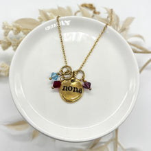 Personalized gold necklace displayed on a small, white ceramic dish. The necklace features a dainty gold chain and a rustic, organic-shaped gold disc hand-stamped with the name "nona" in a lowercase serif font. Three faceted bicone crystal charms in light blue, ruby red, and amethyst purple hang from textured gold rings alongside the pendant. The background is bright white with soft, blurred dried floral accents framing the dish.