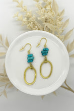 Artisan-style turquoise and gold earrings shown in a flat-lay aesthetic. The jewelry features three stacked turquoise nuggets and an open-oval hammered pendant, evoking a Southwestern or coastal-inspired fashion style.