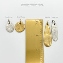 A product size guide shows four artisan charms alongside a brass ruler for scale. The charms feature a hammered, organic texture in silver and gold finishes. Labeled shapes include a "Small Oval" (approx. 0.5"), "Small Round" (approx. 0.5"), "Teardrop" (approx. 1.25"), and "Oval Bar" (approx. 1"). The text at the top reads, "Selection varies by listing." This image helps customers visualize the scale of these jewelry-making su