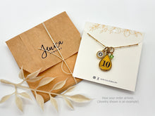 A brown kraft envelope stamped "Jenica JEWELRY" and tied with white twine sits next to a branded display card on a plain white background. The card holds a gold satellite chain necklace featuring three charms: a small initial "O" charm, a yellow resin teardrop charm stamped with the number 10, and a small green crystal. Dried cream foliage accents the left, and lower-right text reads "How your order arrives. (Jewelry shown is an example)".