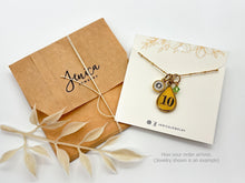 A brown kraft envelope stamped "Jenica JEWELRY" and tied with white twine sits next to a branded display card on a plain white background. The card holds a gold satellite chain necklace featuring three charms: a small initial "O" charm, a yellow resin teardrop charm stamped with the number 10, and a small green crystal. Dried cream foliage accents the left, and lower-right text reads "How your order arrives. (Jewelry shown is an example)".