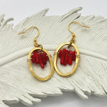 Handmade red coral dangle earrings featuring wire-wrapped vibrant red coral stick beads suspended inside an oval-shaped, textured hammered gold hoop. Displayed on a white decorative feather tray against a clean white background.