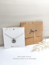 A product photo shows a silver necklace on a white display card next to a brown kraft paper envelope. The necklace features a circular, hand-stamped charm that reads "I love u most" with a heart symbol. The display card has a gold floral design at the top and social media handles at the bottom. The envelope is tied with white twine and features a black cursive logo. A dried white leaf sprig lies in the foreground. Text at the bottom says, "How order arrives. (Jewery shown is an ex