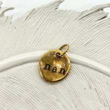 A close-up photograph of a small, organic-shaped gold charm resting on a textured white ceramic feather. The pendant features a hammered, rustic finish and is hand-stamped with the name 'nan' in a black, vintage typewriter-style font. A small, decorative twisted gold jump ring is attached to the top for hanging. The background is a clean, bright white, emphasizing the artisanal detail and warm gold tones of the personalized jewelry piece.