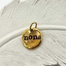 A close-up photograph of a small, organic-shaped gold charm resting on a textured white ceramic feather. The pendant features a hammered, rustic finish and is hand-stamped with the name 'nona' in a black, vintage typewriter-style font. A small, decorative twisted gold jump ring is attached to the top for hanging. The background is a clean, bright white, emphasizing the artisanal detail and warm gold tones of the personalized jewelry piece.