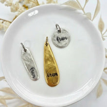 Three hand-stamped "Gram" charms rest on a white ceramic plate, showcasing an organic, hammered texture. The set includes a slender silver oval bar, a long gold teardrop pendant, and a round silver charm, each featuring a rustic artisan finish. Dried white botanicals in the background enhance the neutral, bohemian aesthetic. These versatile pieces are shown with jump rings, ready for use in custom jewelry, bracelets, or as meaningful journal accessories.
