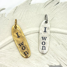 Hand-stamped “I won” charms in gold and silver finishes displayed on a white textured leaf dish. Each elongated oval pendant features rustic, slightly irregular lettering and a handmade look, hanging from small twisted rings. Minimal, neutral background highlights the meaningful, inspirational message and organic design.