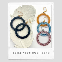 Alt Text:

A flat-lay photograph featuring four pairs of circular resin hoop earrings. Two solid black hoops rest on a white decorative ceramic feather. To the right, three single hoops are stacked vertically: a pearlescent amber-gold, a sparkly glitter blue, and a sparkly glitter pink. The earrings feature various gold and silver hardware, including fishhooks and leverbacks. The image is styled as a Polaroid with the text "BUILD YOUR OWN HOOPS" printed at the bottom against a gray bac