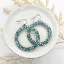 A pair of blue green glitter resin hoop earrings resting in a small white ceramic dish. The hoops are filled with textured silver and muted teal glitter roxs. They are attached to silver-toned fishhook wires with clear rubber safety backs. The dish is styled on a white surface with soft, neutral-toned dried botanicals in the background for a bright, artisan aesthetic.