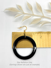 A product photo showcasing a single resin hoop earring on a white background, positioned for scale against a horizontal gold metal ruler. The earring features a thick, high-gloss circular hoop hanging from a gold-toned fishhook wire with a clear rubber stopper. The ruler highlights the hoop's width, which measures 1.5 inches in diameter. A delicate, cream-colored dried botanical sprig is placed in the background, adding a soft, organic touch to the minimalist and clean composition.