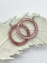A pair of light pink glitter resin hoop earrings displayed on a white, feather-shaped tray. The circular earrings are filled with dense pink and silver sparkling glitter. They feature silver-toned fishhook wires with clear rubber backings. The tray has a textured, ribbed surface mimicking a feather's vanes, all set against a clean, white background.