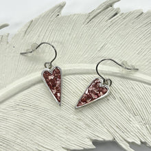 May include: A pair of heart-shaped earrings with a silver-colored frame. The hearts are filled with small, sparkling pink crystals. The earrings hang from silver-colored hooks and are displayed on a white, feather-shaped surface.
