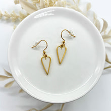 Gemini said
A pair of gold-toned dangle earrings featuring elongated, pointed heart charms. The hearts have a metallic gold border and a smooth, creamy white resin inlay. They are displayed on a white ceramic dish with a subtle textured background of dried botanicals. The earrings are equipped with gold fishhook wires and clear rubber backings.