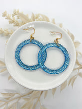A pair of bright blue glitter resin hoop earrings resting in a small white ceramic dish. The hoops are filled with dense, shimmering denim blue glitter. They are attached to gold-toned fishhook wires with clear silicone safety backs. The dish is placed on a white surface with soft, cream-colored dried botanicals blurred in the background, creating a bright, artisan aesthetic.