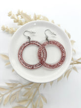 A pair of round pink glitter resin hoop earrings resting in a small white ceramic dish. The earrings feature a vibrant, sparkling rose-pink glitter suspended in clear resin. They are attached to silver-tone fishhook wires with clear silicone backs. The dish is set against a clean white background, accented with soft, neutral-toned dried botanical sprigs, creating a bright and airy aesthetic.