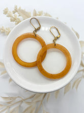 A pair of pearlescent amber resin hoop earrings resting in a small white ceramic dish. The hoops have a warm, golden-orange shimmer with a smooth finish. They are attached to antique gold-toned leverback ear wires. The dish is set against a bright white background with soft, neutral-toned dried botanicals blurred in the background, creating a clean and artisan aesthetic.