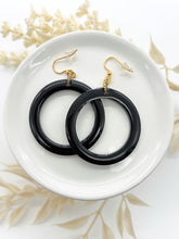 A pair of solid black resin hoop earrings with a high-shine, polished finish resting in a white ceramic dish. They are attached to gold-toned fishhook wires with clear silicone safety backs. The dish is styled on a white surface with soft, neutral-toned dried botanicals in the background for a rustic, artisan feel.