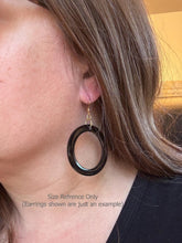 A side-profile photo of a person wearing a large, circular black resin hoop earring to show scale. The earring hangs from a gold fishhook and rests just above the shoulder. Gray text at the bottom reads, "Size Reference Only (Earrings shown are just an example)." The person has long brown hair and is wearing a black shirt against a neutral indoor background.