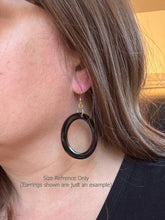 A side-profile photo of a person wearing a large, circular black resin hoop earring to show scale. The earring hangs from a gold fishhook and rests just above the shoulder. Gray text at the bottom reads, "Size Reference Only (Earrings shown are just an example)." The person has long brown hair and is wearing a black shirt against a neutral indoor background.