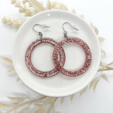 Two pink glitter resin hoop earrings rest on a small white ceramic dish. The earrings are large, circular, and filled with shimmering pink and silver glitter. They are attached to silver-toned fishhook wires with clear plastic backings. The dish sits on a white surface, accented by soft, off-white dried foliage in the background.
