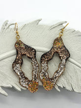 A pair of handmade wavy hoop resin earrings featuring a stunning glitter ombré in shades of brown, champagne, and gold. These boho statement dangles have a unique organic cutout shape and are displayed on a white decorative feather. Designed with gold ear hooks, these lightweight sparkle earrings are perfect for earth-tone jewelry lovers or as a unique handmade gift for her. The shimmering glitter adds a touch of neutral glam to any outfit.